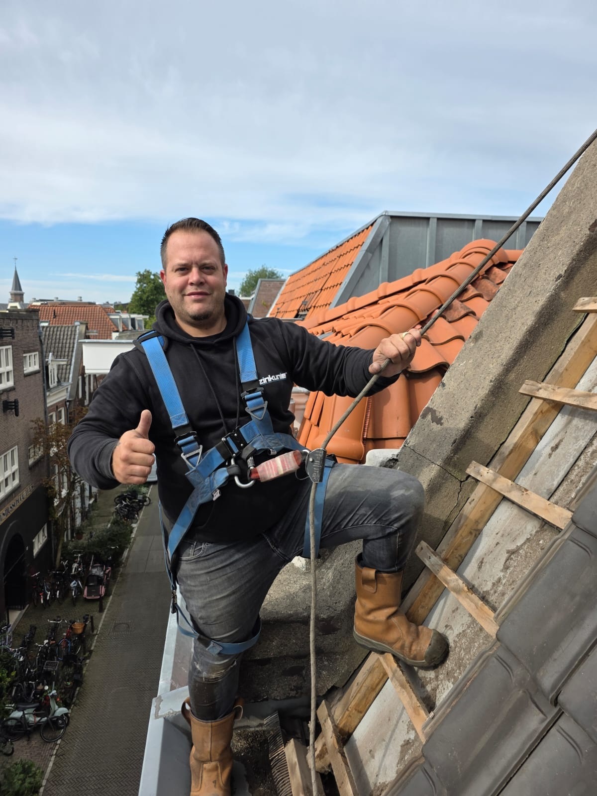 Dakdekker van NH Dak & Lekkages in een veiligheidsharnas op de dakrand, met zijn duim omhoog